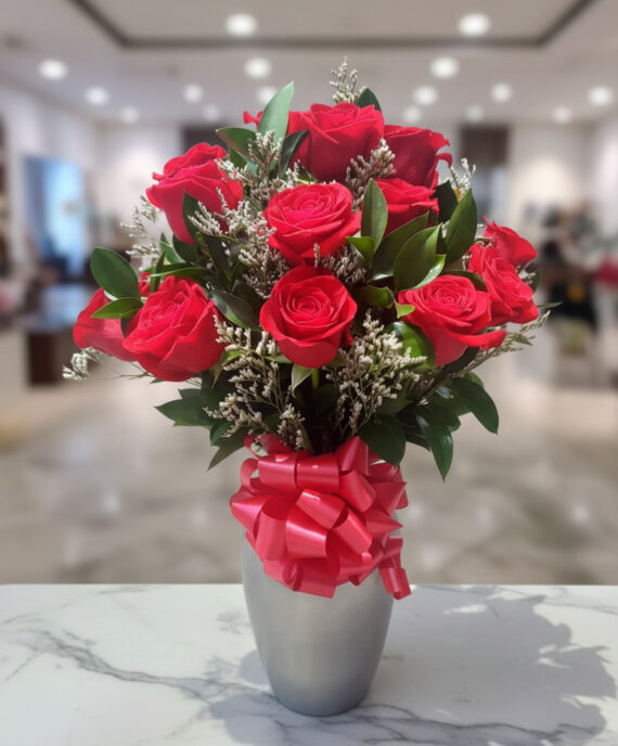 Ramo de rosas rojas con follaje verde y lazo rosa en jarrón plateado sobre mesa de mármol
