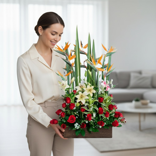 Mujer joven de pie en una sala de estar moderna sosteniendo un arreglo floral rectangular con rosas rojas, lirios blancos y aves del paraíso naranjas