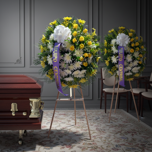 Arreglo floral fúnebre en forma de corona con flores blancas y amarillas, lazo morado con texto Equipo Central, colocado sobre caballete junto a un ataúd de madera barnizada en una sala velatoria elegante