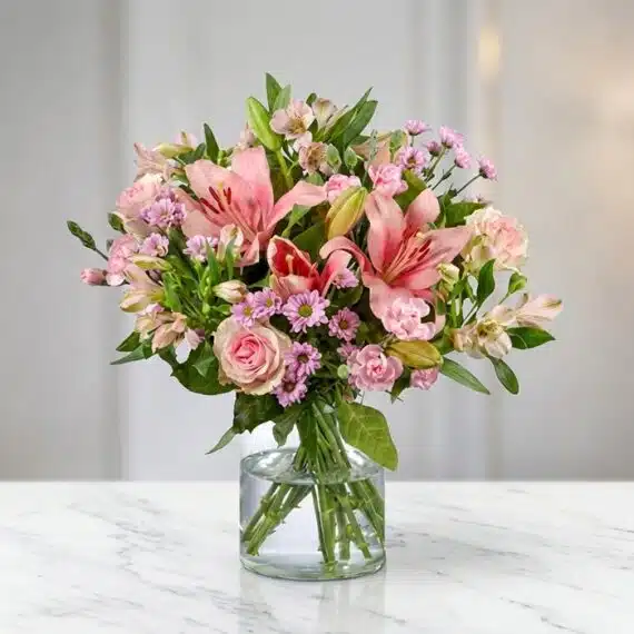 Arreglo Floral ANNA: Delicada Armonía en Rosas y Lirios Arreglo floral ANNA con rosas y lirios rosados, alstroemerias y margaritas, en un jarrón de cristal sobre una mesa de mármol