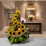 Arreglo floral dorado con girasoles, rosas rojas, uvas, manzanas y peras verdes, sobre una mesa de mármol en un elegante reci