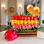 Arreglo floral Escala con rosas rojas, amarillas y rosadas en caja de madera, con globos dorados y lazo rosa, sobre mesa de m