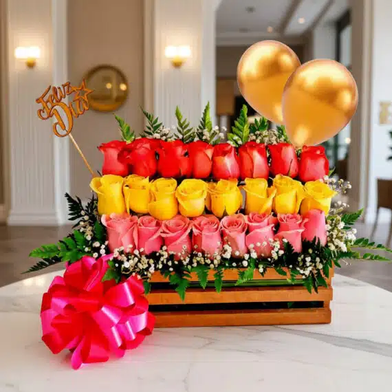 Arreglo Floral Escala: Rosas Rojas, Amarillas y Rosadas con Globos Arreglo floral Escala con rosas rojas, amarillas y rosadas en caja de madera, con globos dorados y lazo rosa, sobre mesa de m