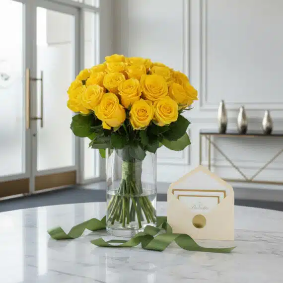 Arreglo floral Evelina con 24 rosas amarillas vibrantes en jarrón de cristal, tarjeta de regalo y cinta verde sobre mesa de m