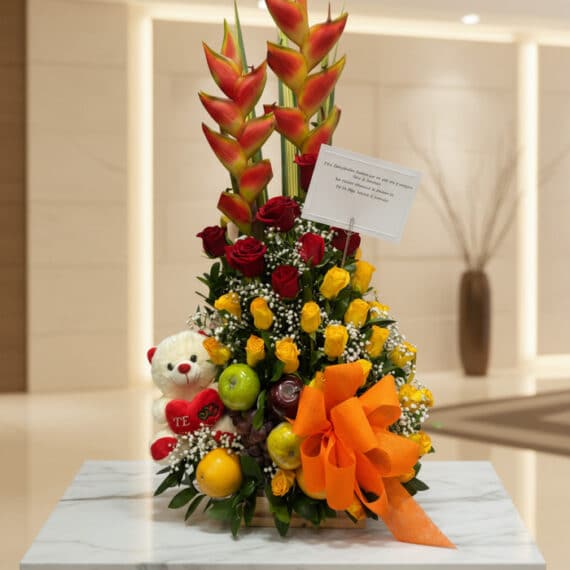 Arreglo floral premium con rosas rojas y amarillas, heliconias, frutas frescas y un lazo naranja, sobre una mesa de mármol en