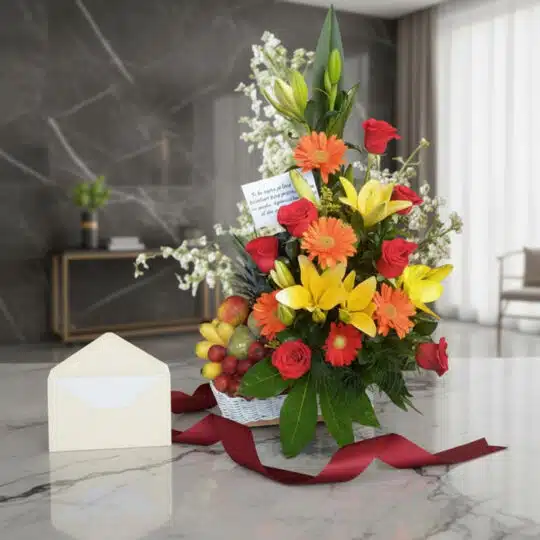 Arreglo floral con rosas rojas, lirios amarillos, gerberas naranjas y frutas variadas en una cesta blanca, sobre una superfic