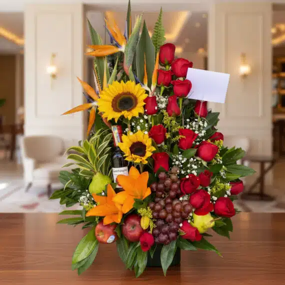 Arreglo Floral con Frutas y Vino: Elegancia para Toda Ocasión Arreglo floral premium con girasoles, rosas rojas, lirios, uvas, manzanas, peras y una botella de vino, sobre una mesa de mad
