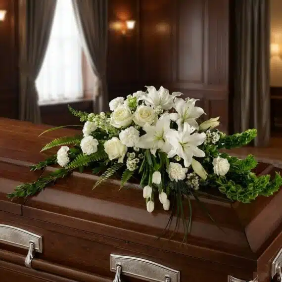Arreglo Fúnebre Cubre Caja Bondad: Elegancia y Respeto en Flores Blancas Arreglo floral fúnebre cubre caja 'Bondad' con lirios, rosas y claveles blancos, sobre un ataúd de madera oscura en una sala