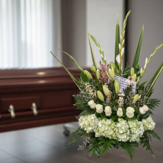 Arreglo floral fúnebre 'Eterno Descanso' con lirios, rosas blancas y hortensias, en sala de velación moderna con ataúd desenf