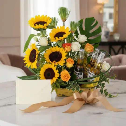 Arreglo floral 'Temis' con girasoles, rosas blancas y naranjas, hojas de monstera y tres botellas de cerveza Corona, presenta