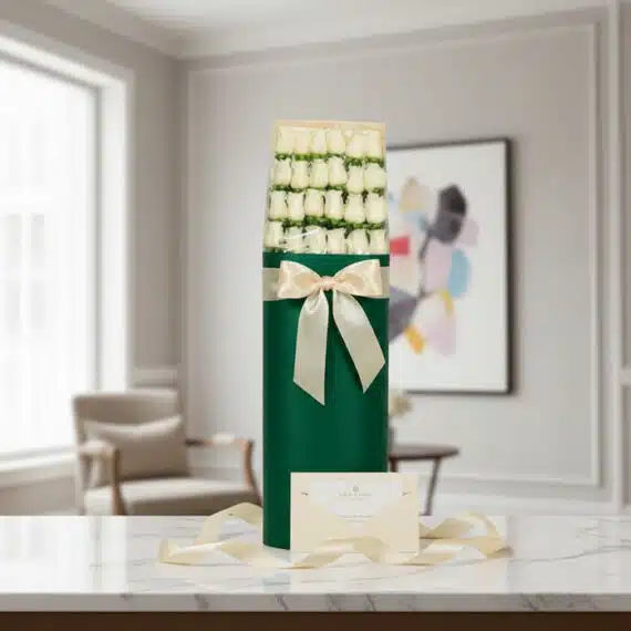 Caja cilíndrica verde con 24 rosas blancas, lazo satinado color crema, tarjeta de regalo y cinta sobre mármol.