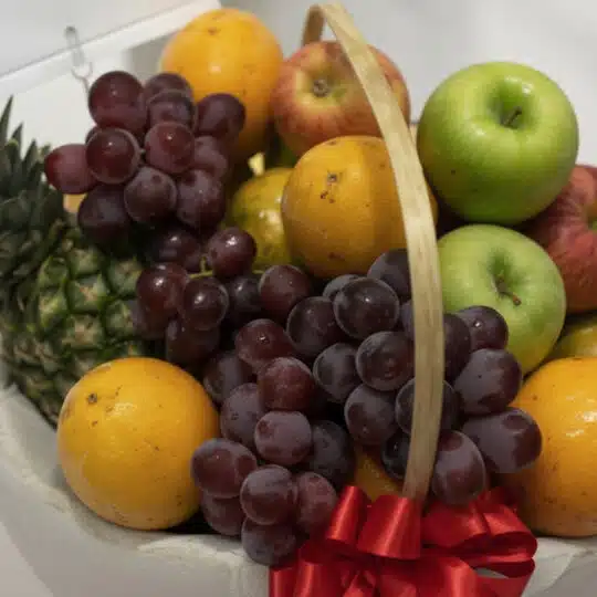 Primer plano de una canasta de frutas gourmet con uvas rojas, manzanas verdes y rojas, naranjas y piña, con un lazo rojo.