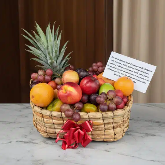 Canasta de Frutas Musca: Frescura y Elegancia para Regalos Especiales Canasta de frutas Musca con piña, uvas, manzanas, naranjas y duraznos, presentada en un entorno elegante con fondo de madera