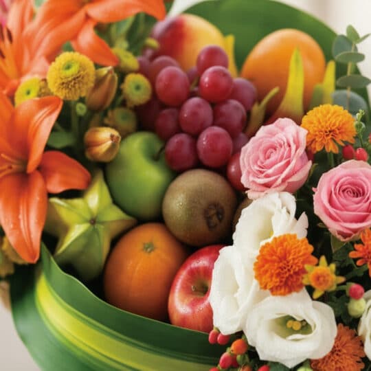 Primer plano extremo de la Cesta Frutal THALIA con lirios naranjas, rosas rosadas, uvas rojas, manzanas, kiwi y carambola, de