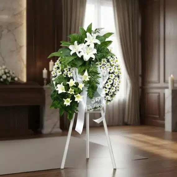 Corona de Homenaje «El Vuelo de Uriel» con Lirios y Margaritas Blancas Corona de homenaje "El Vuelo de Uriel" con lirios blancos, margaritas y follaje verde, sobre un trípode blanco en una sala de