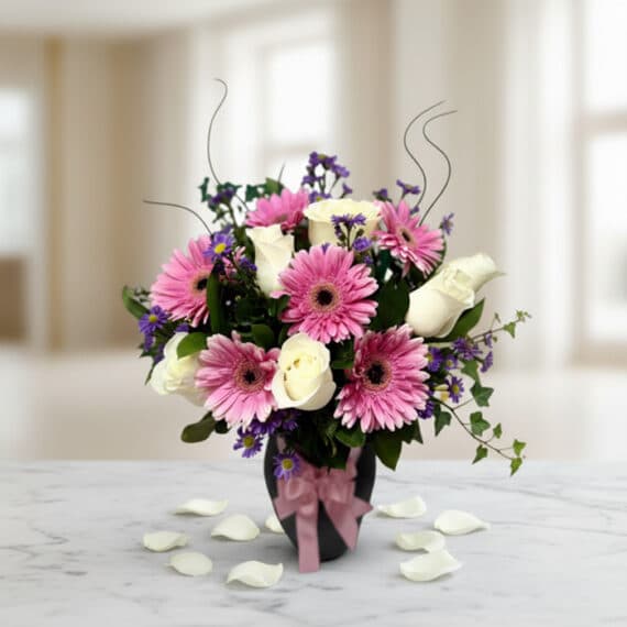 Jarrón Primavera: Elegante Arreglo Floral con Rosas y Gerberas Jarrón Primavera con un elegante arreglo floral de rosas blancas, gerberas rosas y pequeñas flores moradas, sobre una mesa de