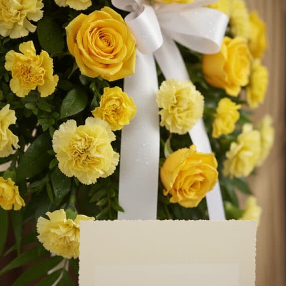 Detalle macro de arreglo fúnebre 'Legado Sereno' con rosas y claveles amarillos, follaje verde, rocío sutil y cinta satinada
