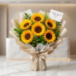 Ramillete de seis girasoles grandes y vibrantes con follaje verde, envuelto en papel coreano beige y blanco con un lazo de sa